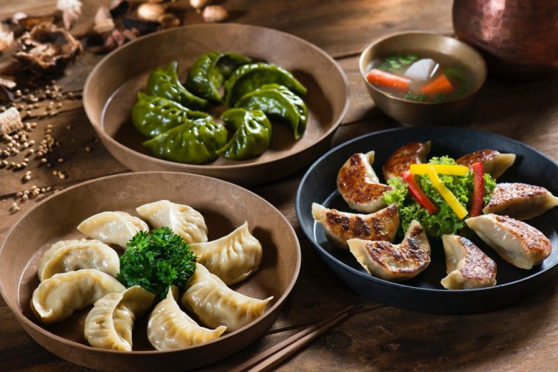 three circular metal plates on a table with crescent shaped steamed dumplings or momos laid out in a circle on each plate around a small pile of greens. What to eat in Nepal. 