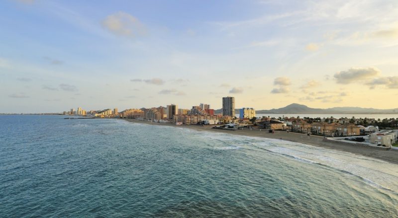 Landscape showing a narrow peninsula with a sandy beach backed by a row of houses and high rise hotels with the sea on either side and mountains on the far side of the bay in the distance. Reasons to Visit Murcia