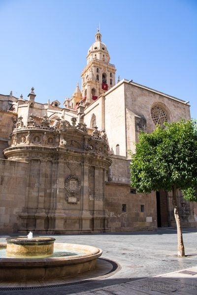 A small circular fountain in an empty square next to a tree with a historic church behind built from beige coloured stone. 