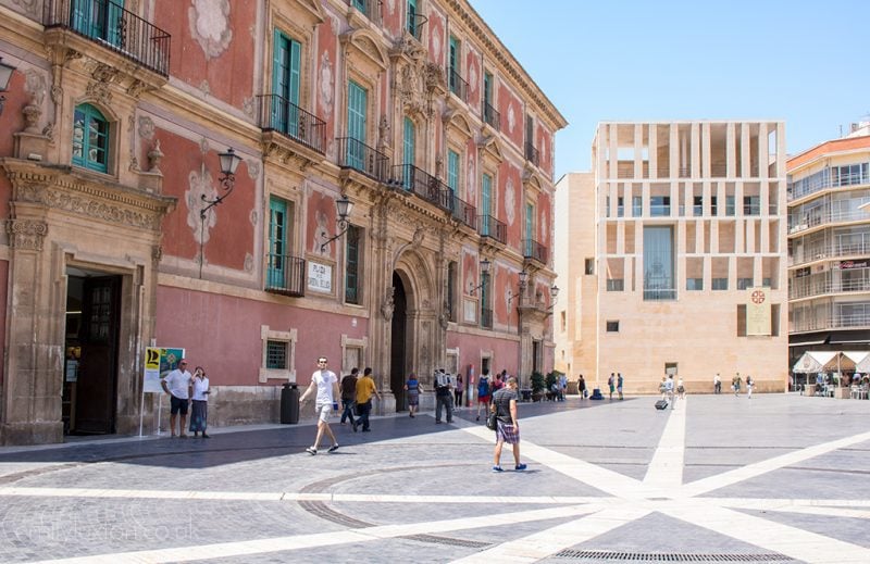 Central plaza in Murcia old town, a large public square with grey paving with a large palace style mansion to the left which has a pink facade, and a modern building behind built from beige stone with many square shaped holes in the facade