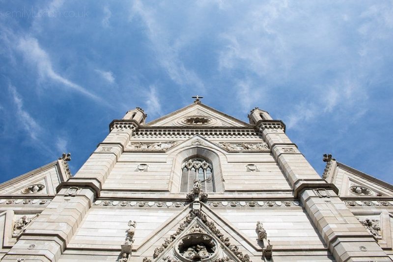 Looking up at the front of Naples cathedral with a triangular roof and a white stone facade against a blue sky