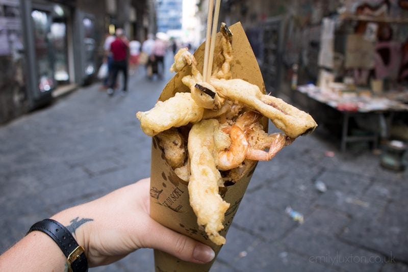 Hand holding a paper cone filled with fried battered vegetables and shrimp in front of a grey flagstone street in naples.