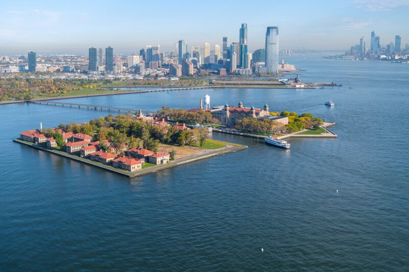 Aerial view of Ellis Island with NYC behind and a ferry arriving at the museum building