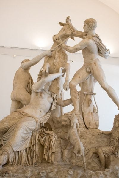 Large white marble statue depicting two nude men and a woman wearing a robe around her lower half wrestling with a large bull inside the Naples Archaeology Museum