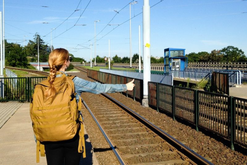 girl wearing a mustard yellow carry on backpack with blonde hair in a lait standing next to a train track making the hitchhiking sign with her thumb out