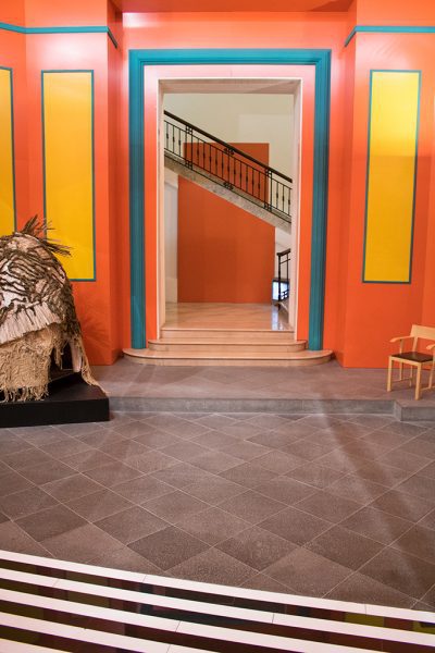 Entrance hall at the MADRE museum with grey tiled floot and an empty doorway in a wall painted bright orange with blue stripes and yellow rectangles