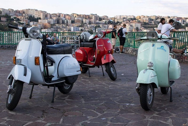 3 vespa mopeds parked on a flagstone courtyard on a viewpoint with a view of Naples city skyline behind. From left to right the vespas are white, red, and pale green. 