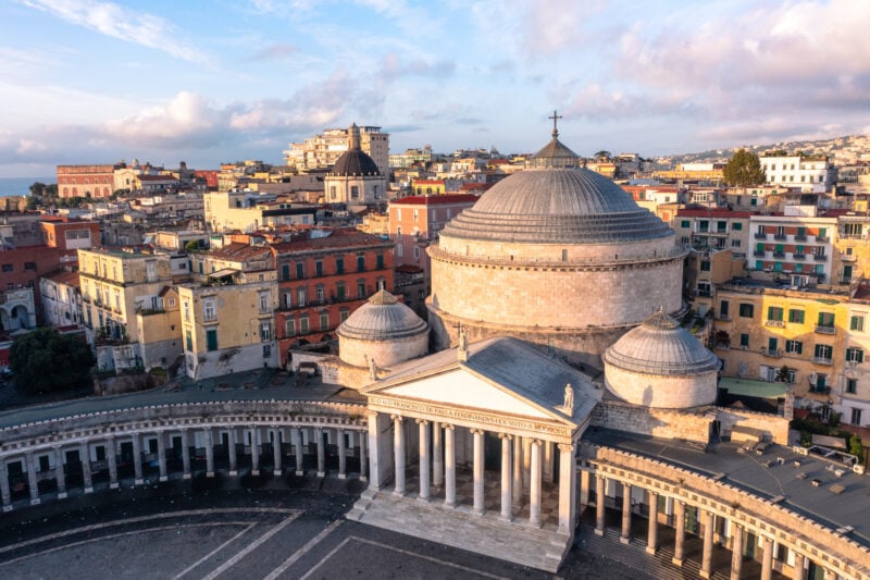 Aerial View of the Basilica Reale Pontificia San Francesco da Paola in the Piazza del Plebiscito in Naples. The basilica is a large circular stone building with a conical roof and a small roman style white temple in front with a long curved covered walkway on either side with collonades. the colourful buildings of naples old town are visible behind. 