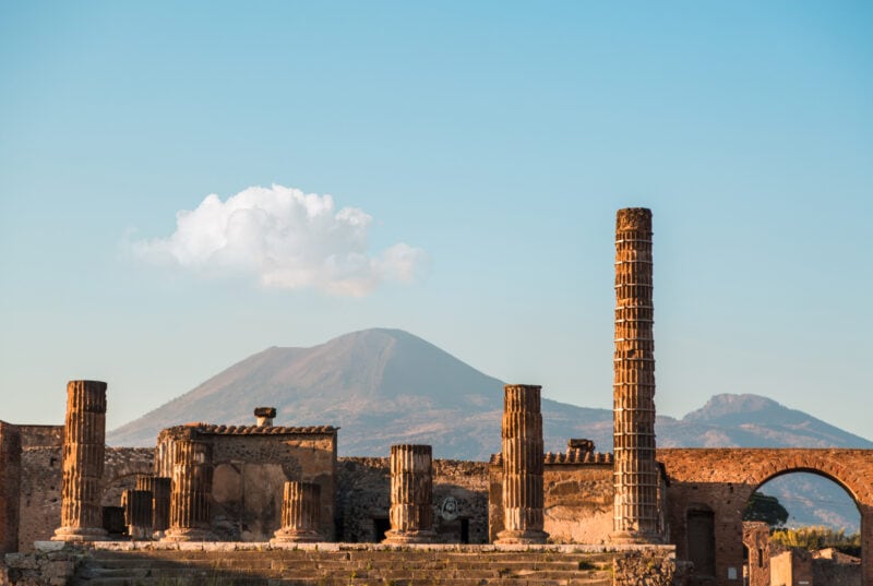 ruined roman buildings with 6 columns in front and a view of Mount Vesuvius behind at Pompeii in Italy