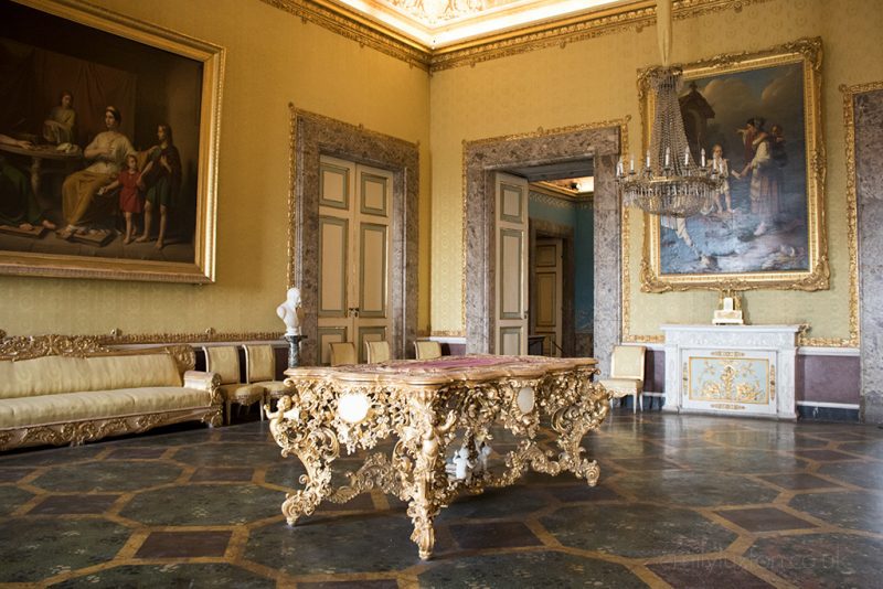 Room inside the Reggia di Caserta with yellow walls with gold trim around the edges and an elaborately carved golden table in the center of the room. 