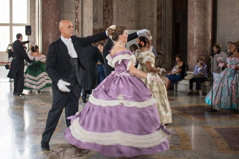 Man in a tuxedo and a woman in a purple ball gown with a large bell skirk and white lace trim dancing in the centre of the ballroom at the Reggia di Caserta with marble floor and marble columns around the sides of the room. There are more couples dancing out of focus behind them.