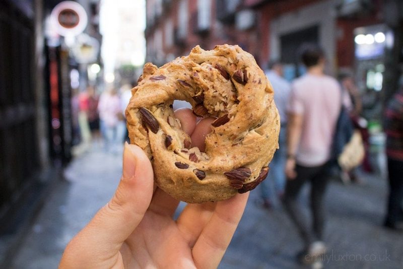 Hand holding a ring shaped biscuit with raisins in and a hole in the middle in front of a crowded out of focus street in Naples
