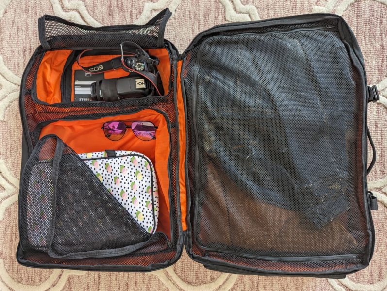 flat lay photo of a black backpack open with an orange interoior, one side is closed with a black mesh cover and the other side contains a camera, pink sunglasses, and a white wash bag