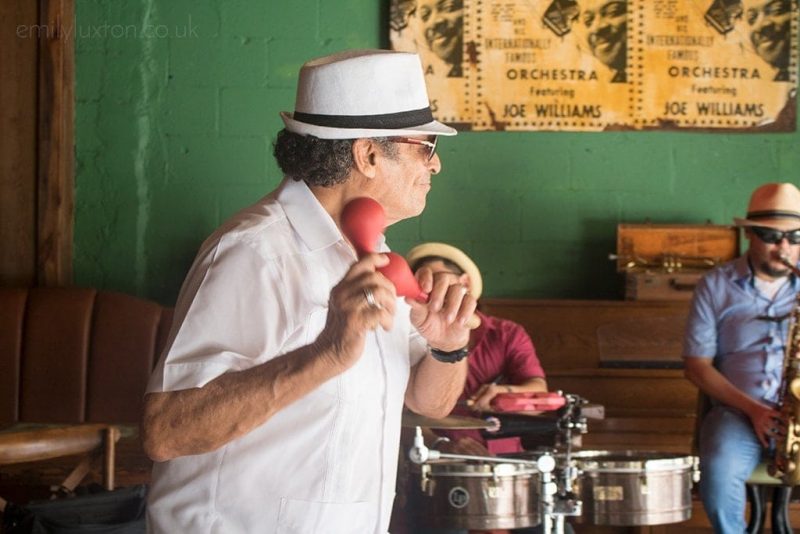 a man in a white shirt and white trilby hat dancing and shaking maracas in a bar with a green wall behind him and 2 members of the the band out of focus playing steel drums and trumpet. 
