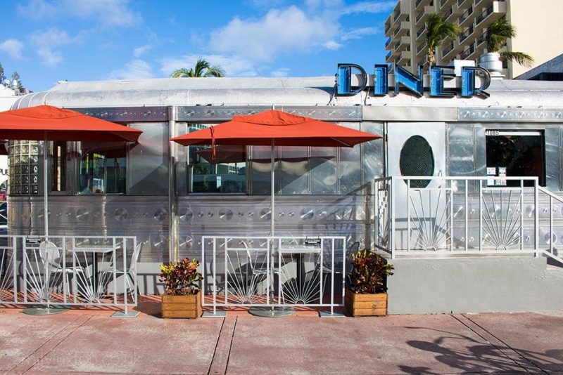 front of the 11th street diner in miami, a silver metal train car with windows and a blue sign on top that says diner, with two grey tables out front with red umbrellas. 