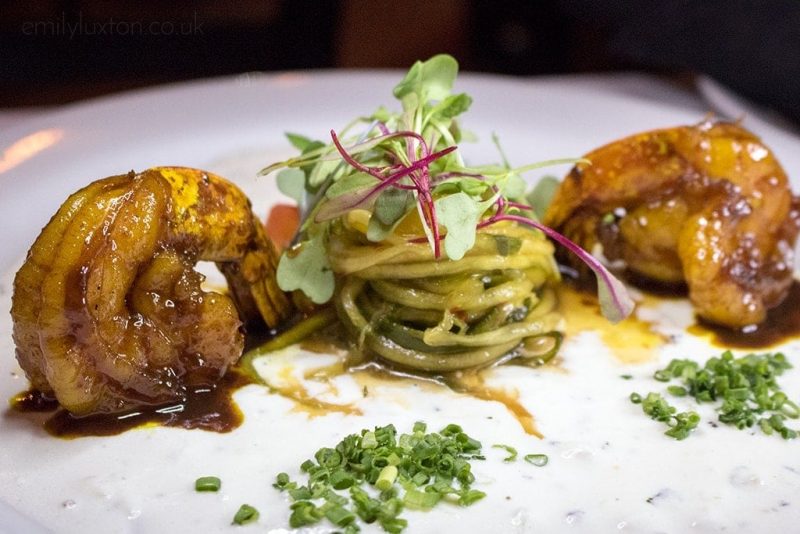 close up of a plate of food with two shrimps marinated in a yellow sauce on either side of a small twist of spaghetti topped with greens