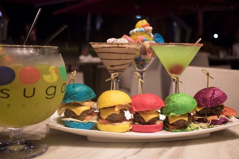 5 tiny burgers with different colour buns left to right: blue, yellow, red, green, purple. the burgers are lined up on a white plate with a large yellow cocktail in front and two small cone glasses containing cocktails behind.