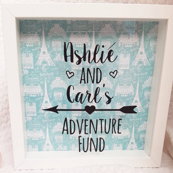 Close up of a wooden box with a clear plastic front printed with the words Ashlie and Carl's adventure fund.