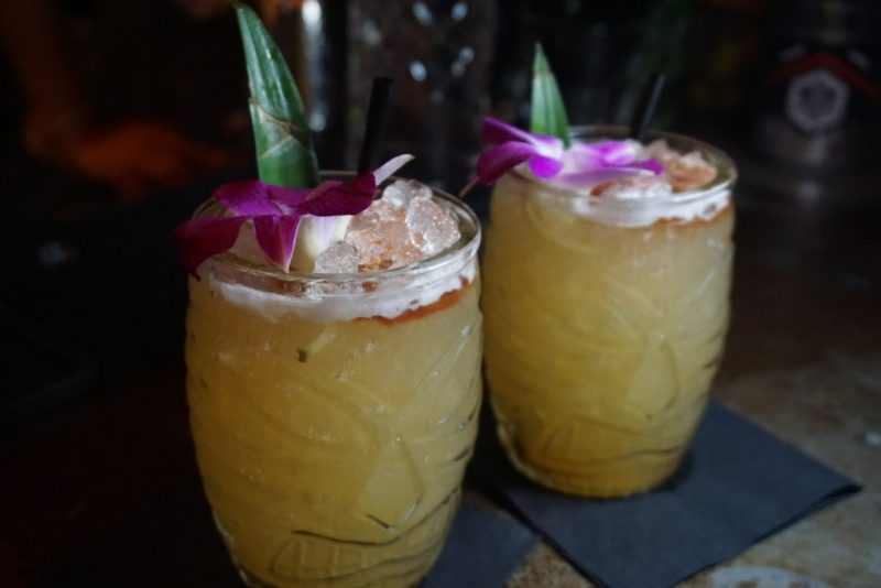 close up of two yellow cocktails in tiki glasses topped with a purple flower and a peace of palm leaf 