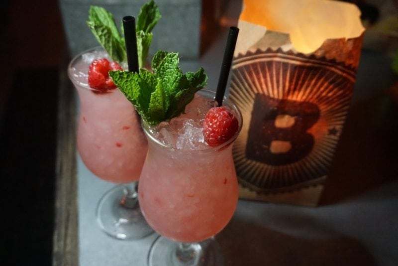close up of two pale pink cocktails in tall glasses with a raspberry on top and a sprig on mint and black straws. at a bar in miami. 