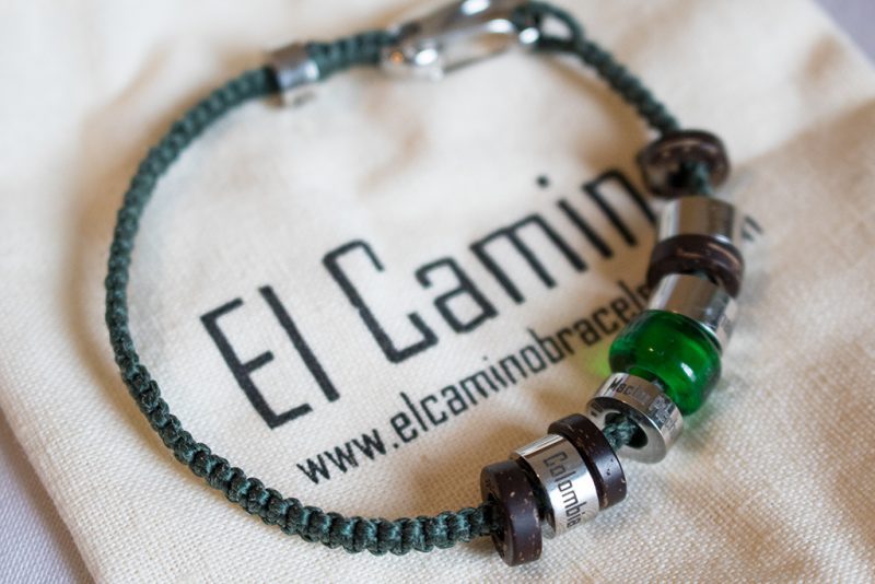Close up of a bracelet made from green rope with wooden and silver metal beads on it. The bracelet is on a small canvas bag with the brand name El Camino printed in black.