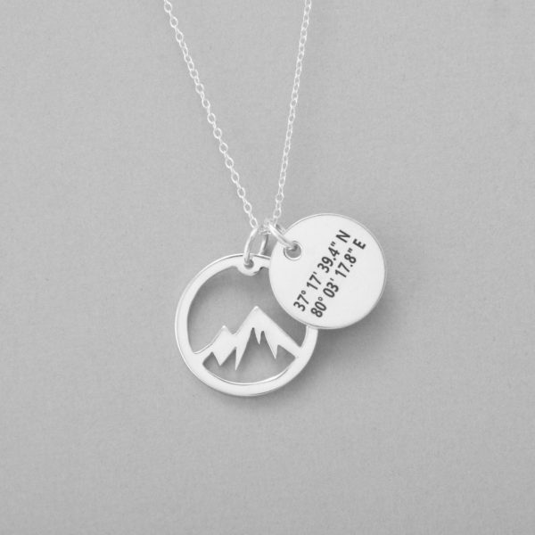 Close up of a silver necklace with two circular pendants, one in the shape of a mountain and one engraved with coordinates.