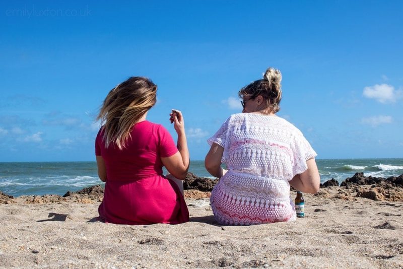 Two blonde women sitting on a sandy beach with their backs to the camera looking at the sea on a sunny but windy day. The girl on the left is wearing a red dress and her hair is blowing in the wind, the girl on the right is wearing a white crochet kaftan. Fun Things to do in Hutchinson Island Florida