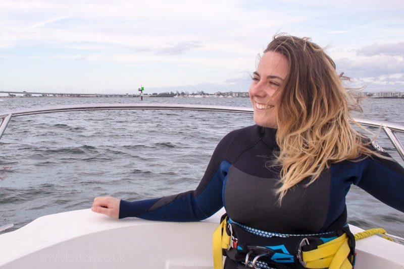 emily wearing a black wet suit with blue and grey trim. Her long blonde ombre hair is down and blowing in the wind. She is sitting on the edge of a white boat with the grey sea behind her and smiling as she look saway to the left. fun things to do in hutchinson island florida.