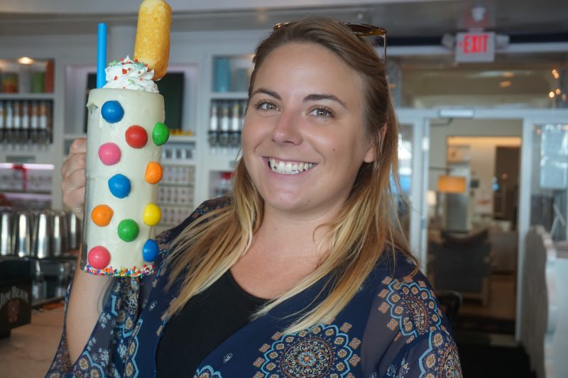 Emily holding a oversized glass with a while milkshake dotted with large colourful gumballs and topped with cream, sprinkles, and a twinkie with a blue straw next to it. Emily is wearing a blue patterned shawl and smiling at the camera, with only head and shoulders in the frame and an out of focus restaurant interior behind. Sugar Factory restaurant in Miami