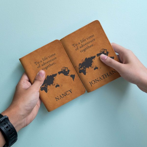 Close up of two hands each holding a brown leather passport cover, both covers have a map engraved in black and the following text written above: To a life time of adventure together.