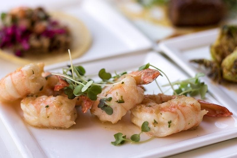 close up of 4 shrimps on a white square plate topped with melted butter and greens. new orleans food and drink