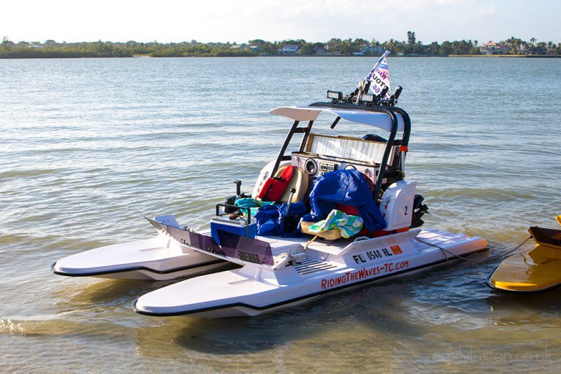 a small white motor catamaran with a flag on the back and red sign at the bottom saying ridingthewaves-tc. the boat is on shallow water in the sea with a green strip of land in the distnace behind.