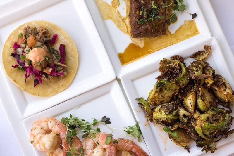 flatlay of several square white plates of food at a new orleans restaurant - top left has an open taco with pulled pork, greens and red cabbage. top right has a piece of meat with greens and a thin brown sauce. bottom left has 4 shrimps with greens and bottom right has fried cabbage with a butter sauce/ 