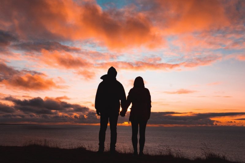 Silhouette of a man and woman holding hands in front of the sea with a sunset sky above. Unique valentine's gifts for travel lovers