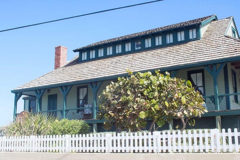 an old fashioned american house with cream cladding, turquoise wooden pillars and a grey slate roof with a white picket fence in front and a blue sky overhead