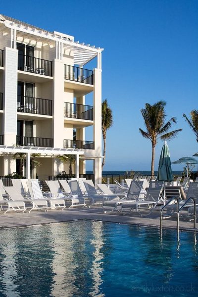 exterior of a white hotel with black balcony railings with a rectangular pool in front on a sunny day with an empty blue sky in Hutchinson Island Florida