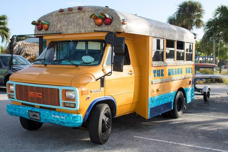 a yellow USA public school bus which has been converted to add a patterned grey roof and bright blue trim around the bottom. the sign on the bus reads The Ohana Bus. There are some paddleboards attached to the back of the bus, which is parked in a car park with palm trees behind. 