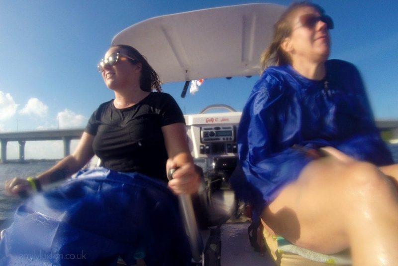 Two women slightly out of focus because of water on the camera lens, sitting on a small motorboat. the girl on the left is wearing a black dress and using a silver metal stick to drive the boat. the girl on the right is wearing a bright blue poncho. they are both soaking wet. Things to do in Hutchinson Island Florida