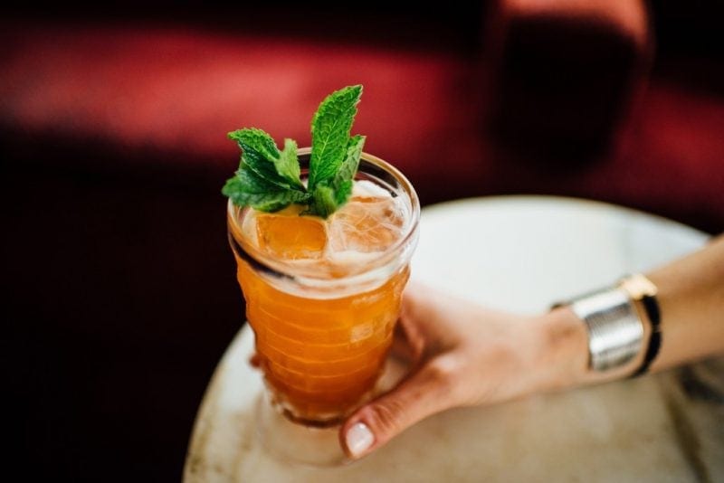 close up of a hand holding an orange cocktail in a tall glass with ice and a sprig of mint on a white table against a dark red background