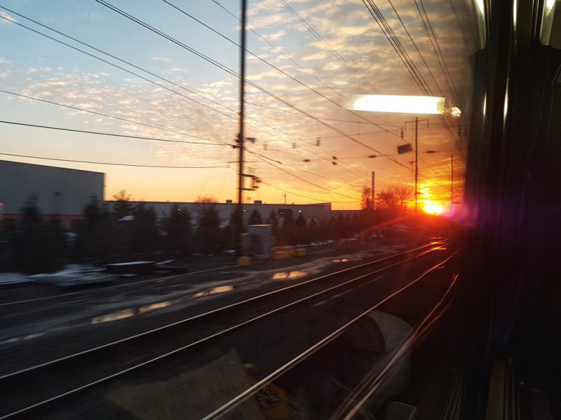 Looking out a train window at a sunset with train tracks and telegraph wires and a few white industrial buildings. Amtrak Crescent review. 