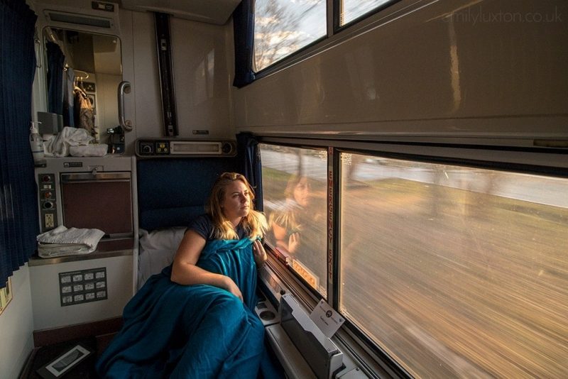 Blonde girl wrapped in a blue blanket on a blue chair in a train cabin looking out the window with the ground out of focus rushing by outside. Amtrak Crescent review
