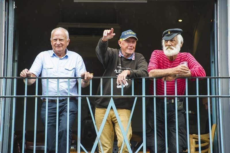 looking up at a balcony with three elderly men leaning on the railings, the man on the right has a white beard and a is wearing an old fashioned sailing cap, the central man is wearing a blue cap and pointing at something, the man on the left is wearing a light blue shirt and facing the camera. 