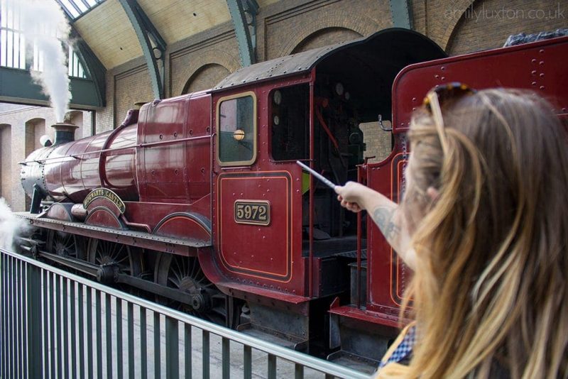 the back of emily's head with one arm raised to point a wand at the scarlet red steam train engine of the Hogwarts Express at Universal Studios Florida