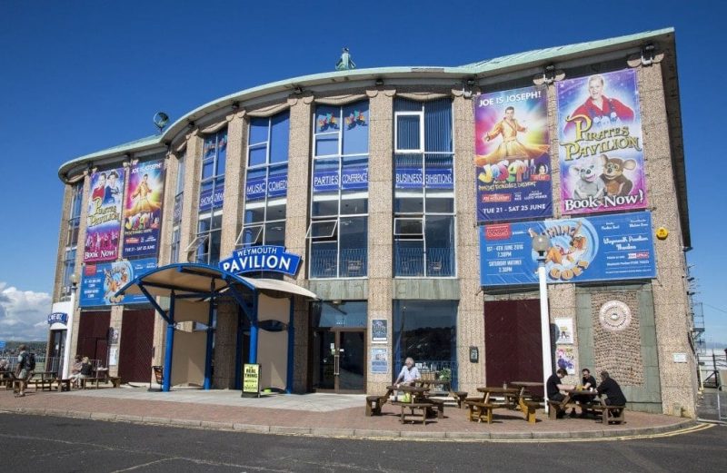 exterior of a large art deco style building of grey stone with a green metal roof, the blue sign above the door reads "weymouth paviliion"