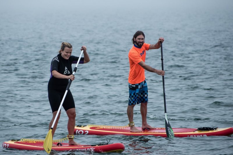 a man and woman standing up on red paddleboards on the sea, the woman is wearing a black rash vest and black shorts and the man has a dark beard and is wearing blue shirts and an orange t shirt.