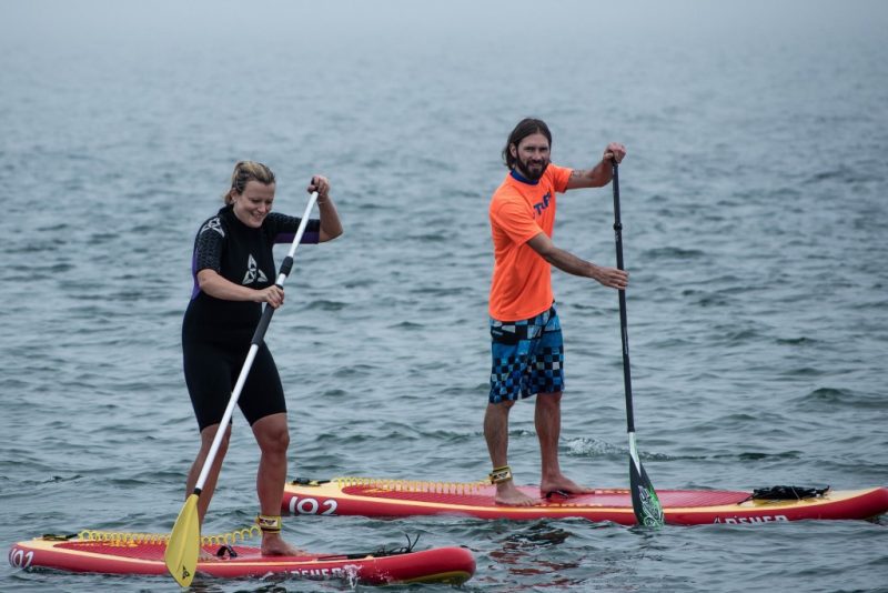 a man and woman standing up on red paddleboards on the sea, the woman is wearing a black rash vest and black shorts and the man has a dark beard and is wearing blue shirts and an orange t shirt. 