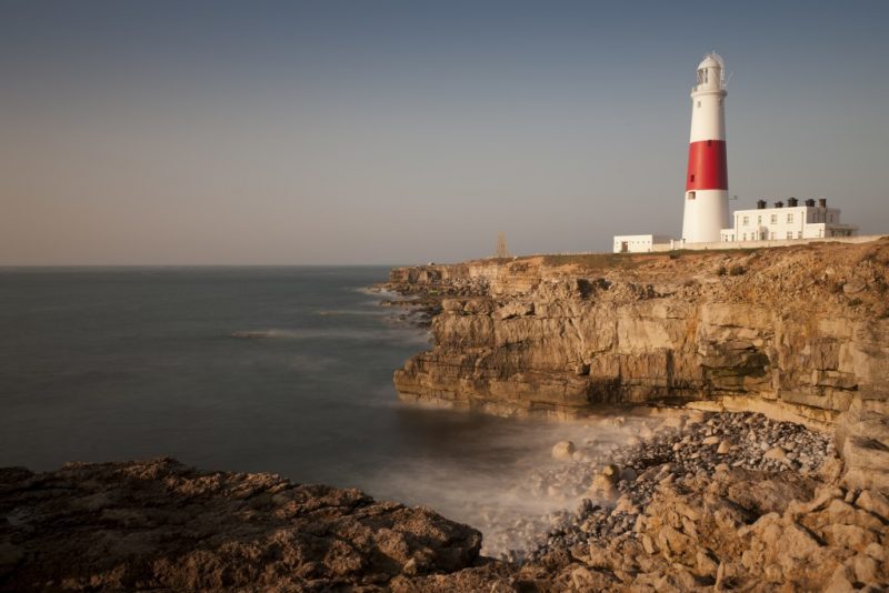 White lighthouse with a red stripe around the middle on a low rocky cliff edge next to the sea at Portland Bill