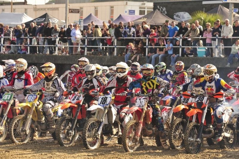 dense crowd of motocross racers on bikes on a sandy beach getting ready to start a race all wearing helmets and goggles, there is a big crowd of specators on the esplanade behind watching.