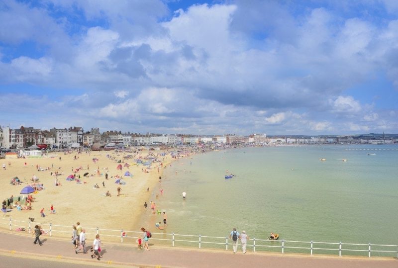 looking down towards a wide sandy beach full of people on a sunny day, with the esplanade visible behind lined by terraced georgian townhouses. 50 Unique Things to do in Weymouth