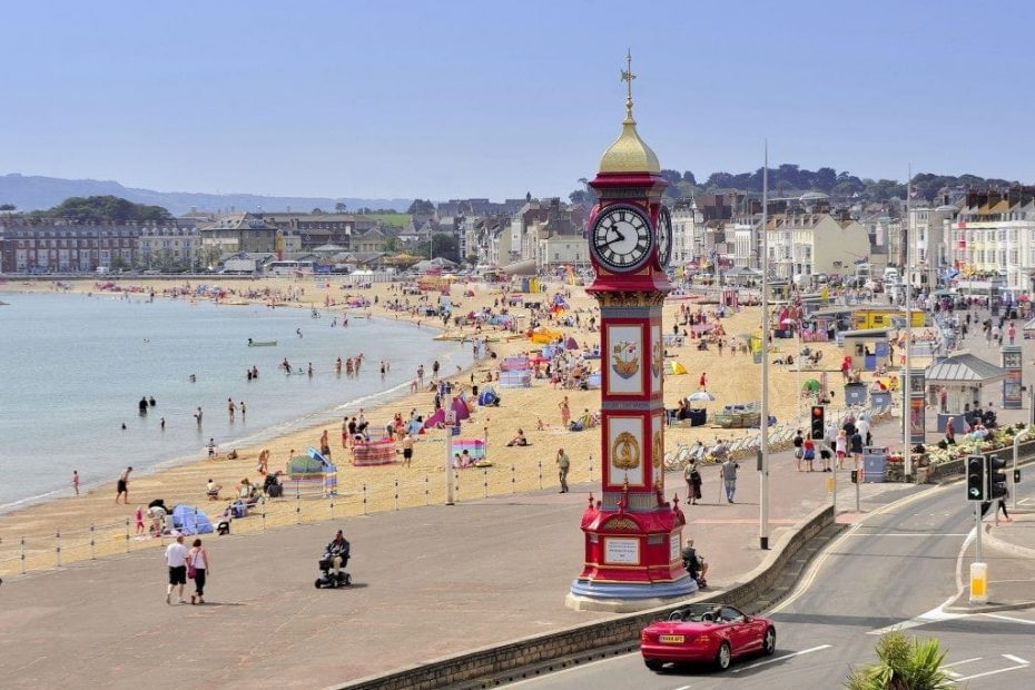 Red clock tower with a golden roof on a wide esplanade next to a sandy beach in Weymouth with a red sports car driving past